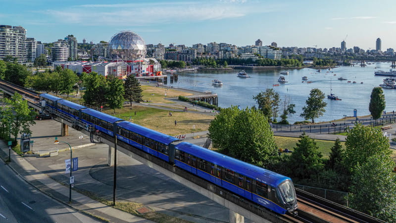 New Mark V train travels along the waterfront with a city skyline in the background.