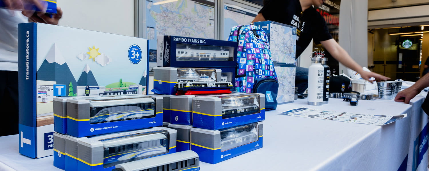 A display table showcases various TransLink store merchandise, including trains, a SeaBus, a backpack, a water bottle, and other souvenirs.