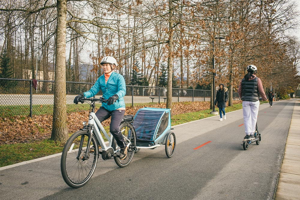 A woman riding an electric bike on a bike path while towing a children's bike trailer.