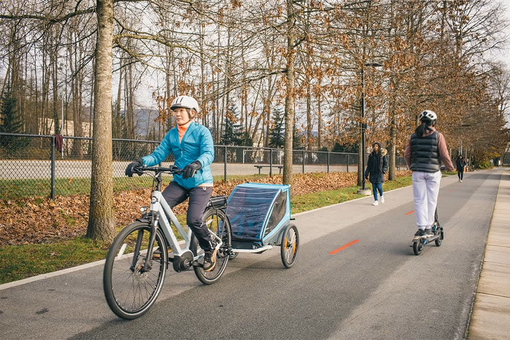 A woman riding an electric bike on a bike path while towing a children's bike trailer.